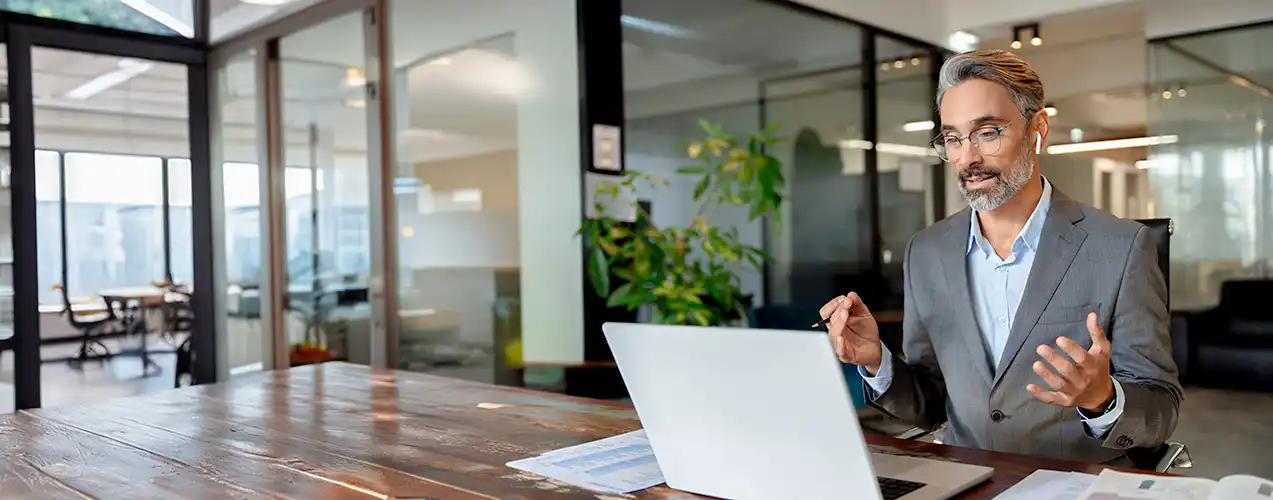 Geschäftsführer kommuniziert via Microsoft Teams mit seinen Mitarbeitenden, die im Homeoffice arbeiten. Im Hintergrund ein leeres Geschäftsbüro.