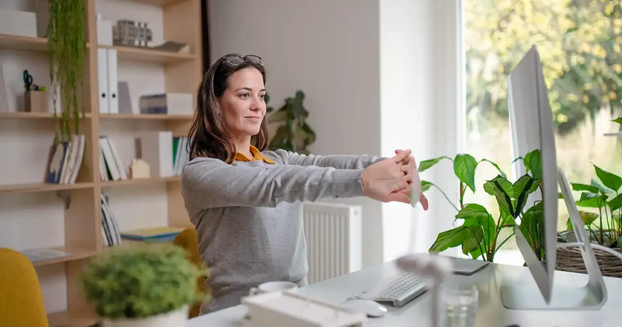 Mitarbeiterin im Homeoffice macht bei einer Pause Dehnungsübungen am Computer.